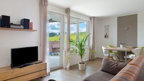 Ein Wohnzimmer mit Fernseher, einer Couch, einem Esstisch mit Stühlen und einem großen Fenster mit Vorhängen.