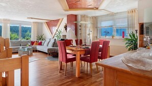 Een ruime woonkamer met een eettafel, rode stoelen, een bank en uitzicht op een bos door grote ramen.