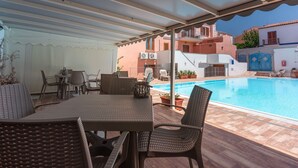 Un área de estar al aire libre con techo, mobiliario de mimbre, piscina y edificios al fondo.