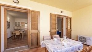 Un comedor con una mesa dispuesta para dos, contraventanas de madera en las puertas y una cocina visible a través de un vano.