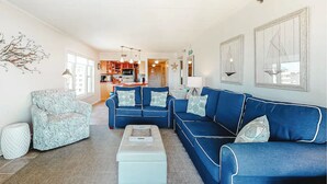 Ein Wohnzimmer mit blauem Sofagarnitur, einem gemusterten Sessel und einem Couchtisch. Im Hintergrund ist eine Küche mit Holzschränken und einer Hängeleuchte zu sehen.