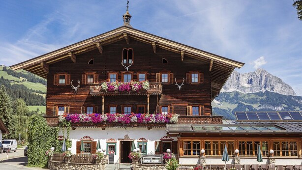 Tipico chalet di legno con balconi, facciata in pietra e montagne sullo sfondo.