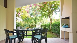 Eine Terrasse mit einem runden Tisch und Stühlen, einem Grill und Blick auf einen Garten mit Bäumen und Sträuchern.