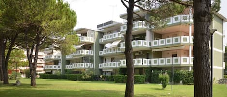 A multi-story residential building with balconies, surrounded by trees and a grassy area.
