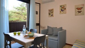 A dining area with a table set for two, a grey sofa, and a window with a view of trees. - Cozy studio apartment for 3 people (Bibione)