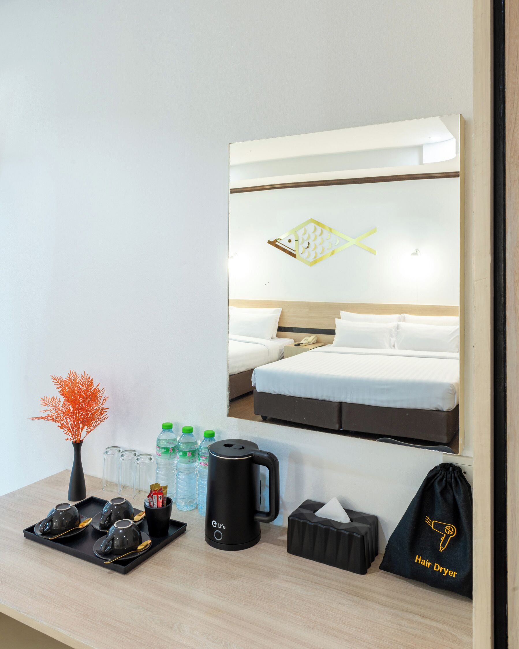 A hotel room with a bed, a bedside table with a vase, water bottles, a kettle, and a hair dryer bag.