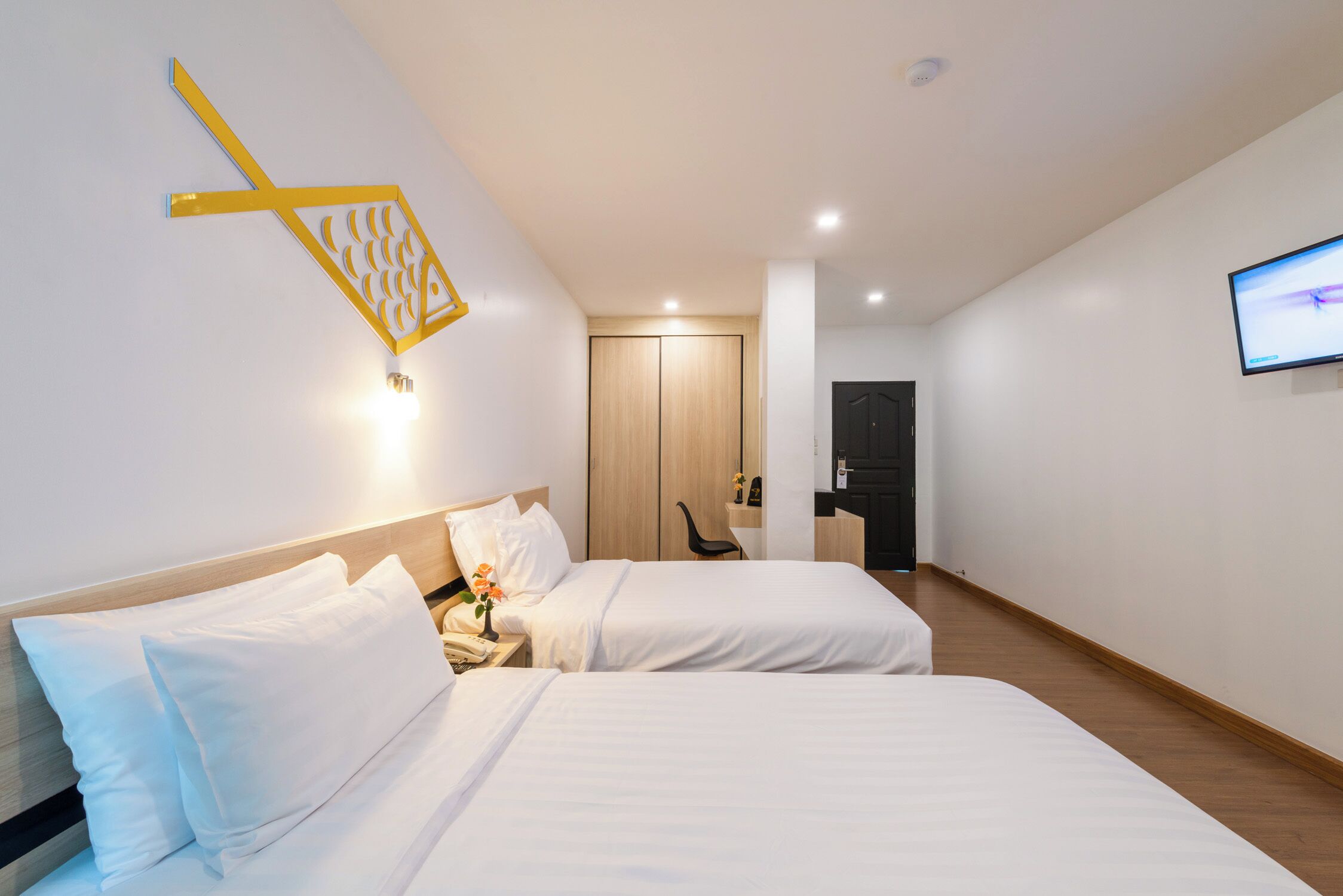 A hotel room with two beds, a wooden wardrobe, a desk with a chair, a TV mounted on the wall, and a unique yellow wall decoration.