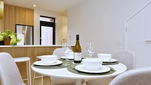 A dining area with a round table set for four, white plates, bowls, and wine glasses. A bottle of wine is on the table. The room has a modern design with light-colored walls and wooden accents. - Modern City Apartment with Style and Comfort (Christchurch)