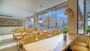 A dining area with wicker furniture, large windows, and a view of pyramids. - La Boutique Hôtel - Vue Des Pyramides (Giza)