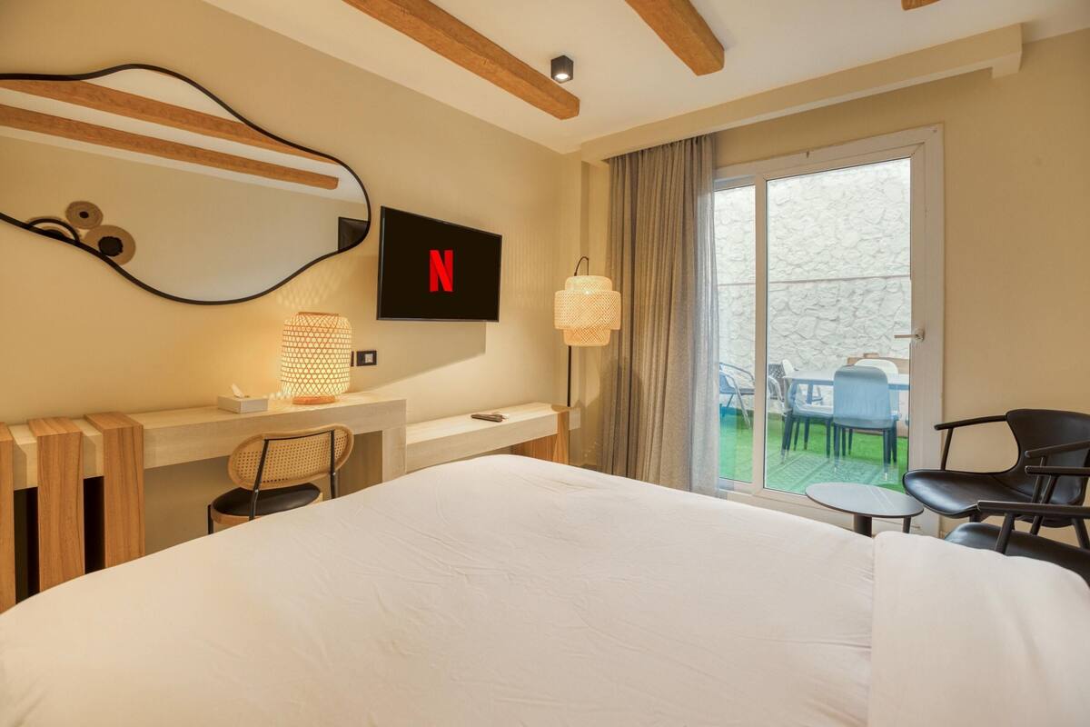 A hotel room with a large bed, a desk with a chair, a TV displaying the Netflix logo, a lamp, and a view of the outdoors through a sliding door.