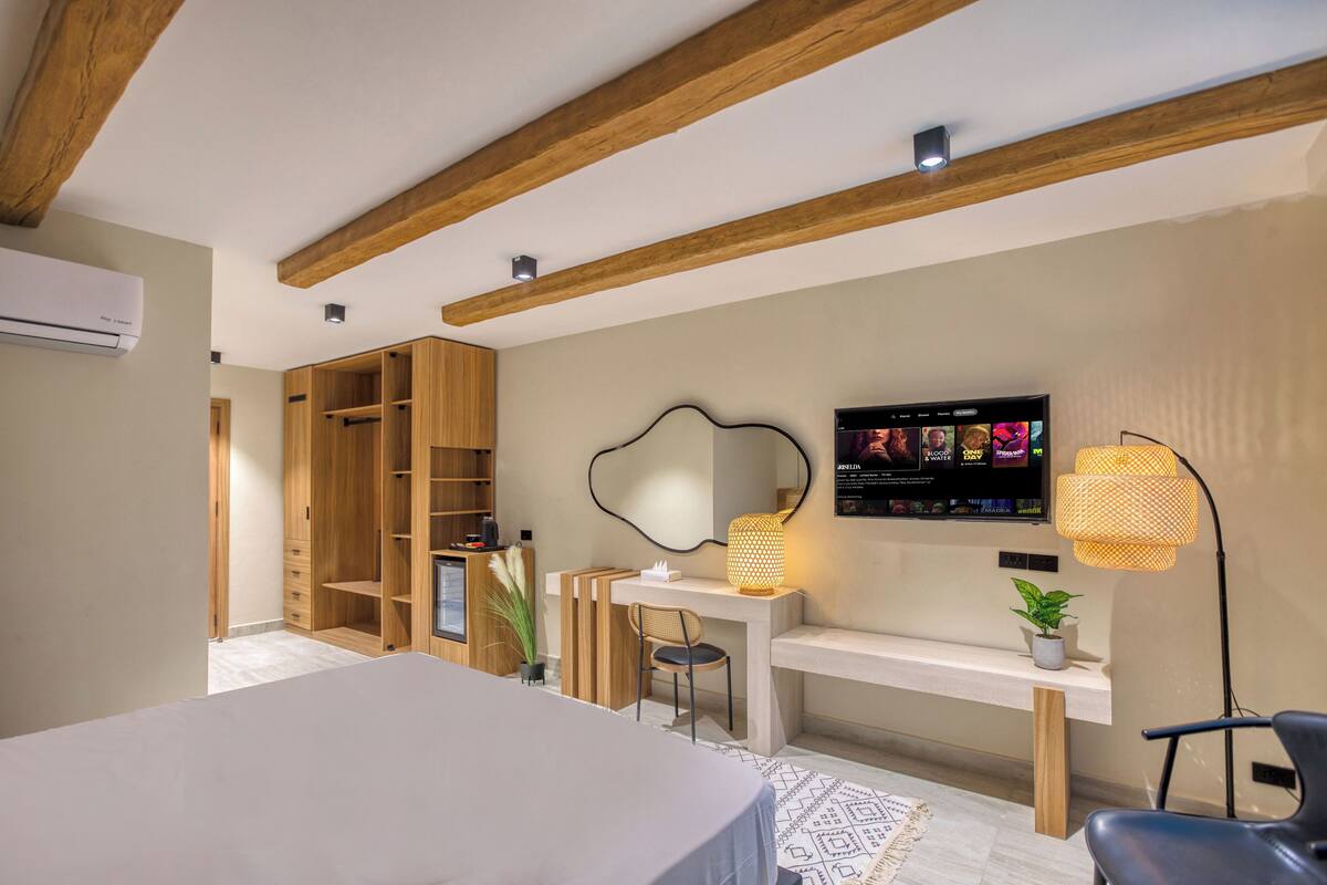 A modern bedroom with a wooden ceiling, a TV mounted on the wall, a desk with a lamp, and a chair.