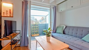 A modern living room with a grey sofa, a wooden coffee table, a TV on a stand, and a balcony with a view of buildings. - 1 bedroom nice apartment in Carolles (Carolles)