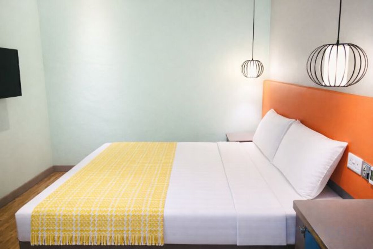 A bed with white bedding and a yellow throw, a nightstand, a wall-mounted TV, and a pendant light.
