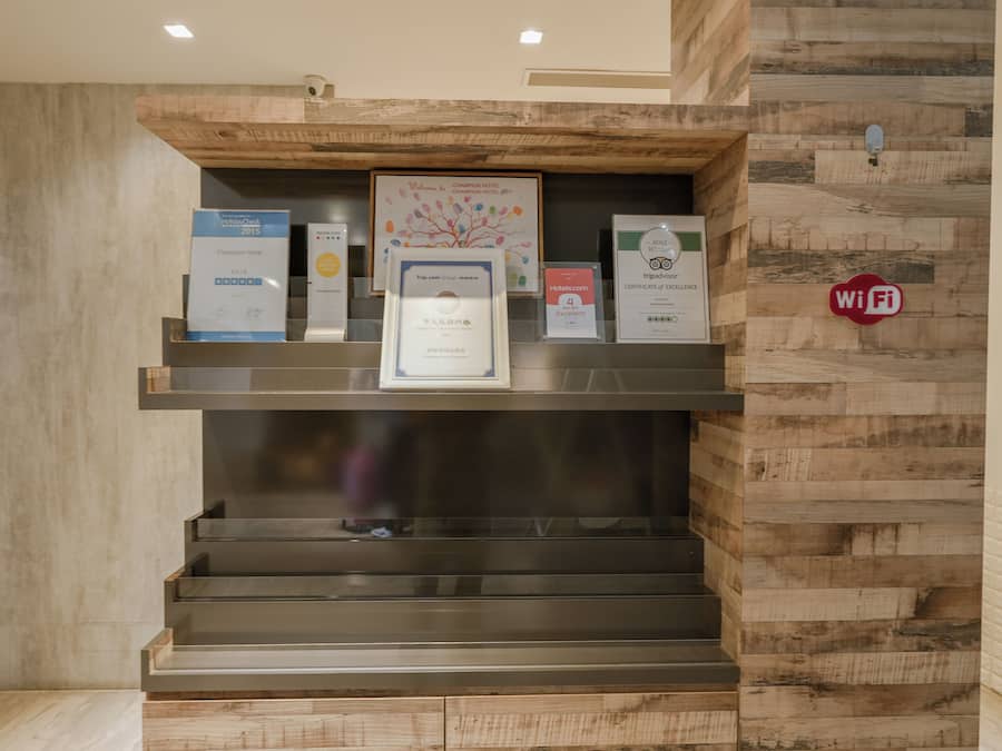 A reception area with a wooden wall and a metal staircase. There are several awards and certificates displayed on the wall, including a TripAdvisor certificate of excellence and a 4-star rating from Hotels.com.
