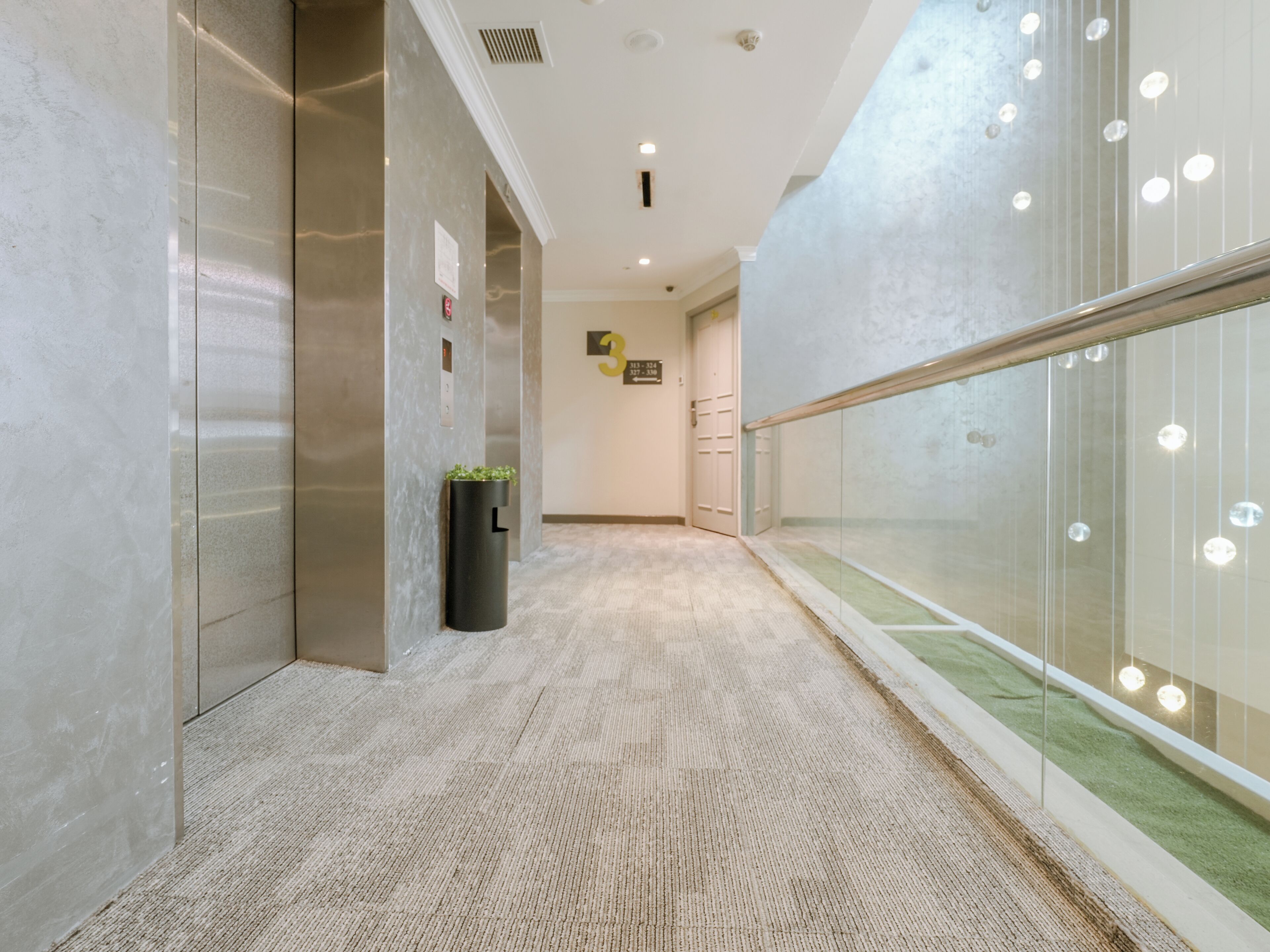 A modern hallway with a marble floor, a black trash bin with a plant, and a glass wall with hanging lights.