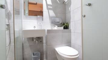 Casa de banho moderna com chuveiro envidraçado, lavatório branco, sanita e uma pequena planta num vaso no balcão.