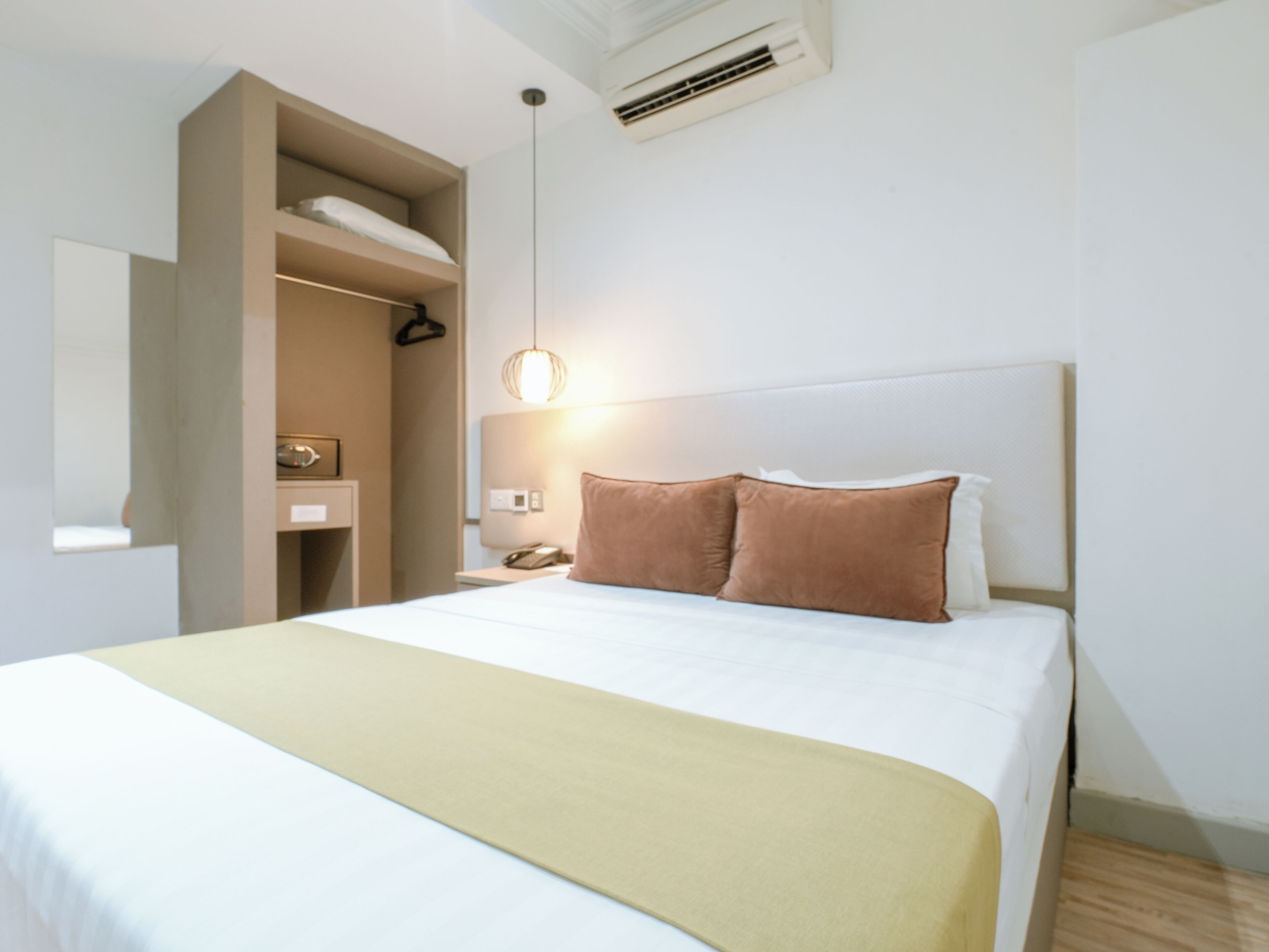 A modern hotel room with a large bed, a bedside table with a lamp, a wardrobe, and a hanging light fixture.