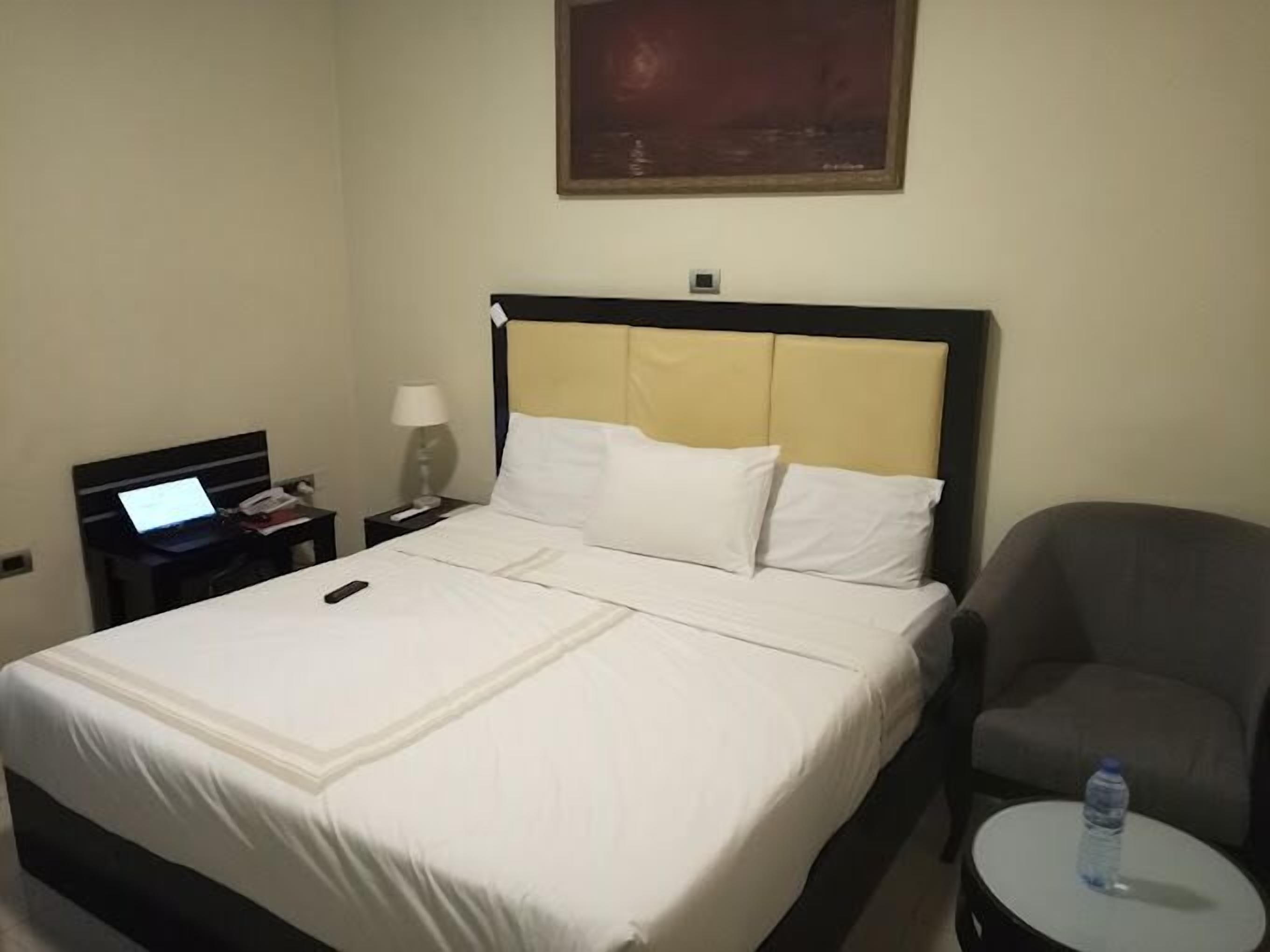 A hotel room with a bed, a desk with a laptop, a chair, and a small table with a water bottle.