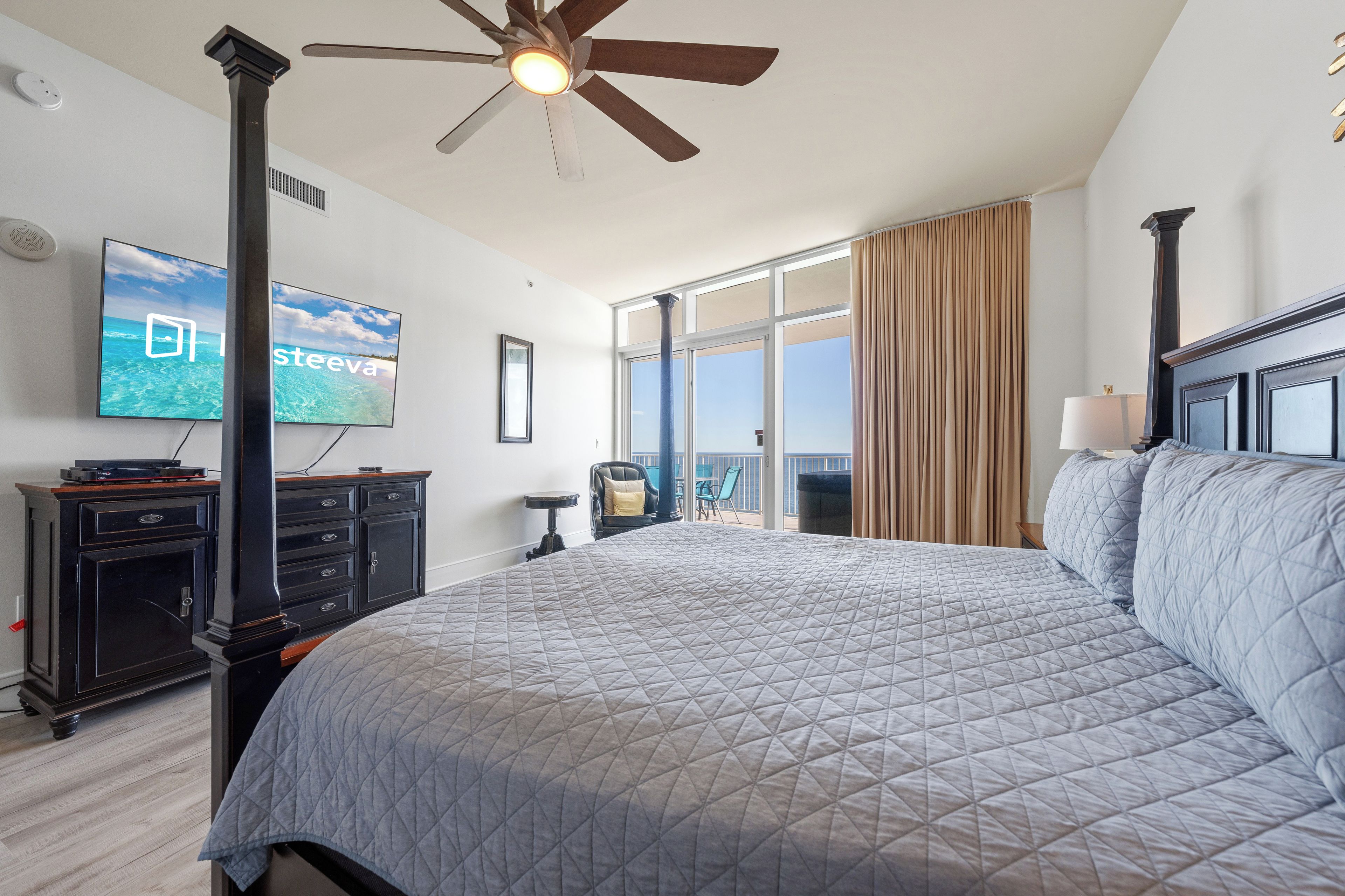 Una camera d'albergo con un letto grande, una ventola a soffitto, una TV fissata al muro e vista sull'oceano dalla finestra.