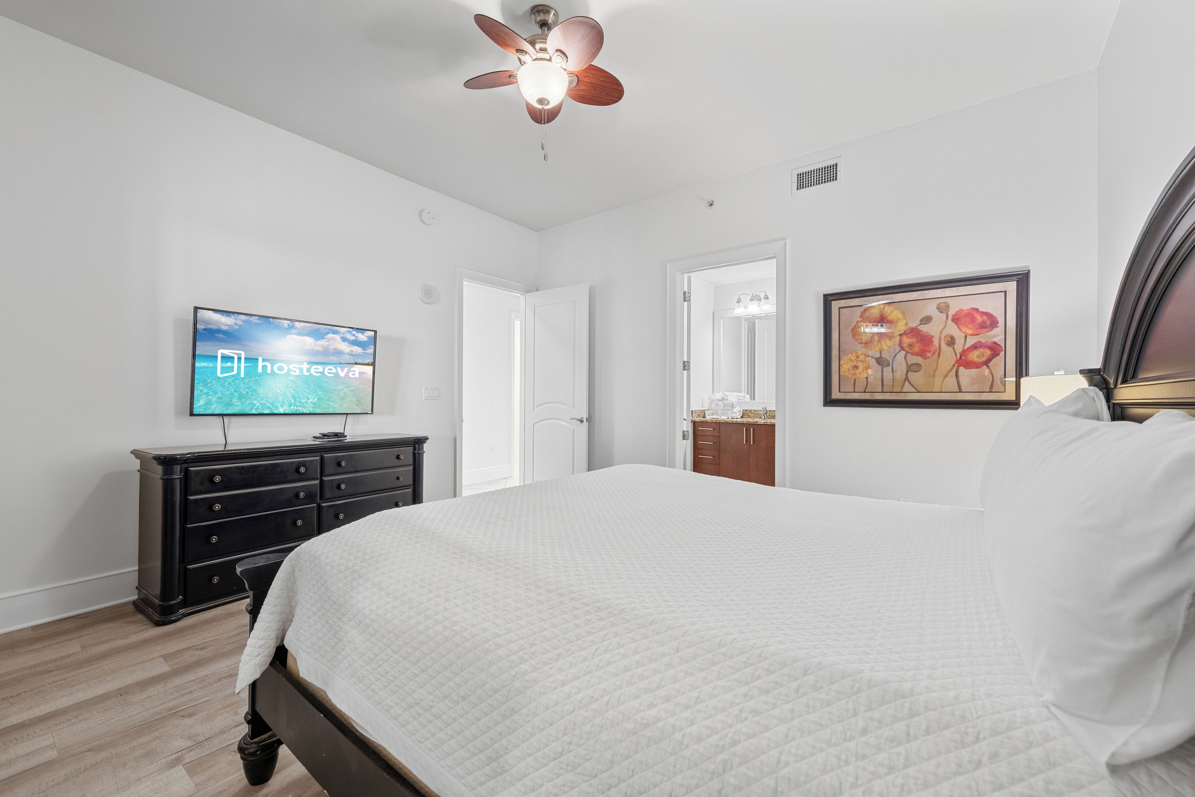 A bedroom with a bed, a television, a ceiling fan, a dresser, and a framed picture on the wall.