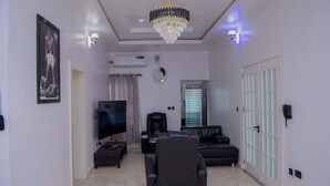 A modern living room with a flat-screen TV, a black leather sofa, a recliner, and a chandelier. - Heineken Beach House  (Lagos)