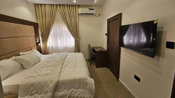 A hotel room with a bed, a desk, a chair, a TV mounted on the wall, and an air conditioning unit. - Heineken Beach House  (Lagos)