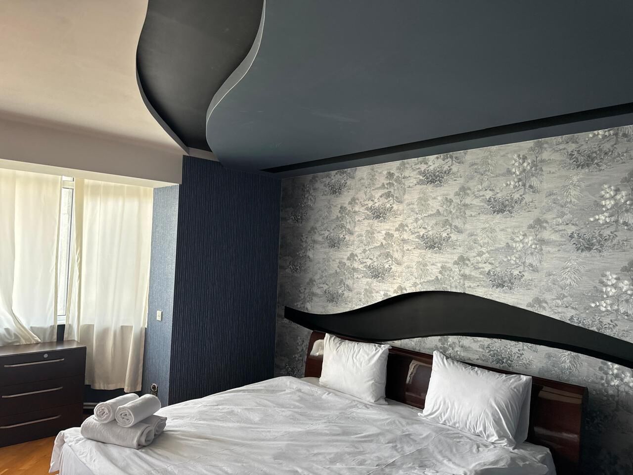 A modern bedroom with a large bed, dark headboard, and a decorative wall with floral wallpaper.