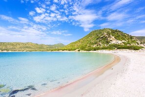 A beach with clear turquoise water, white sand, and a green hill in the background.