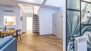 Un apartamento moderno con suelo de madera, zona de comedor con mesa y sillas, cocina con ventana, escalera y radiador con papel tapiz tropical.