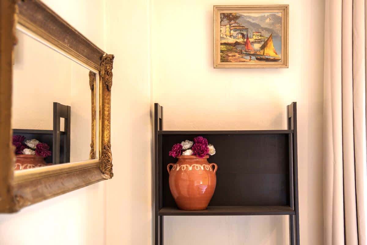 En hall med en stor, utsmyckad spegel, en inramad målning som föreställer ett kustlandskap och en hylla med en terrakottafärgad vas med blommor.