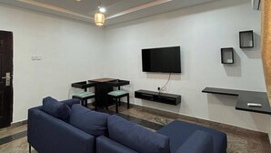 Un salon moderne avec un canapé d’angle, un téléviseur à écran plat fixé au mur, une table à manger avec des chaises et un éclairage encastré au plafond.