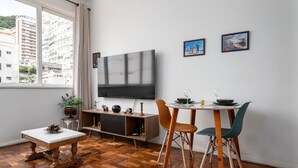 Un salón moderno con una televisión grande, suelo de madera, una mesa de comedor con sillas y vistas a un paisaje urbano a través de la ventana.