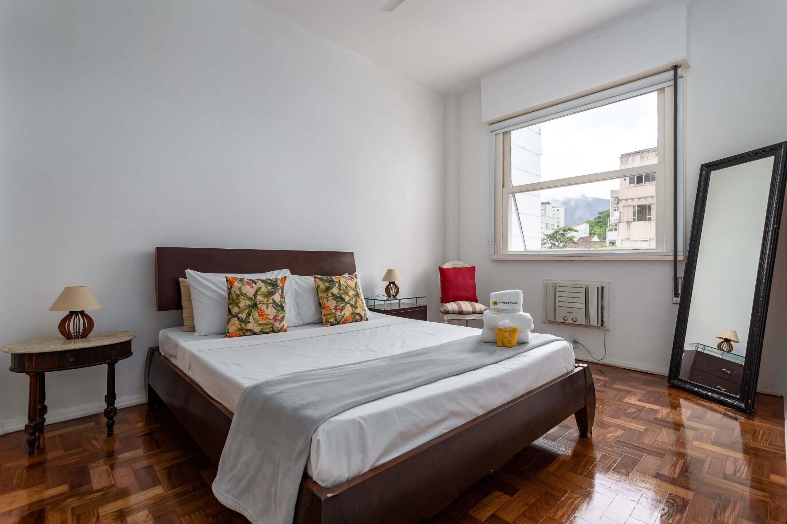A bedroom with a wooden bed, white bedding, a nightstand with a lamp, a red chair, and a window with a view of buildings.
