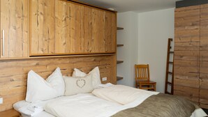 Un dormitorio con cabecera de madera, cama con ropa blanca, armario de madera y una silla de madera.