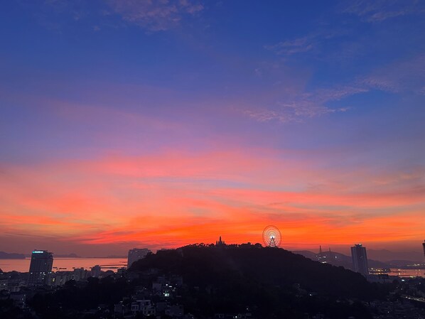 A cityscape at sunset with a prominent Ferris wheel on a hill, buildings in the foreground, and a body of water in the distance. - MonHomestay Ha Long-Amazing view balcony (Hong Gai)
