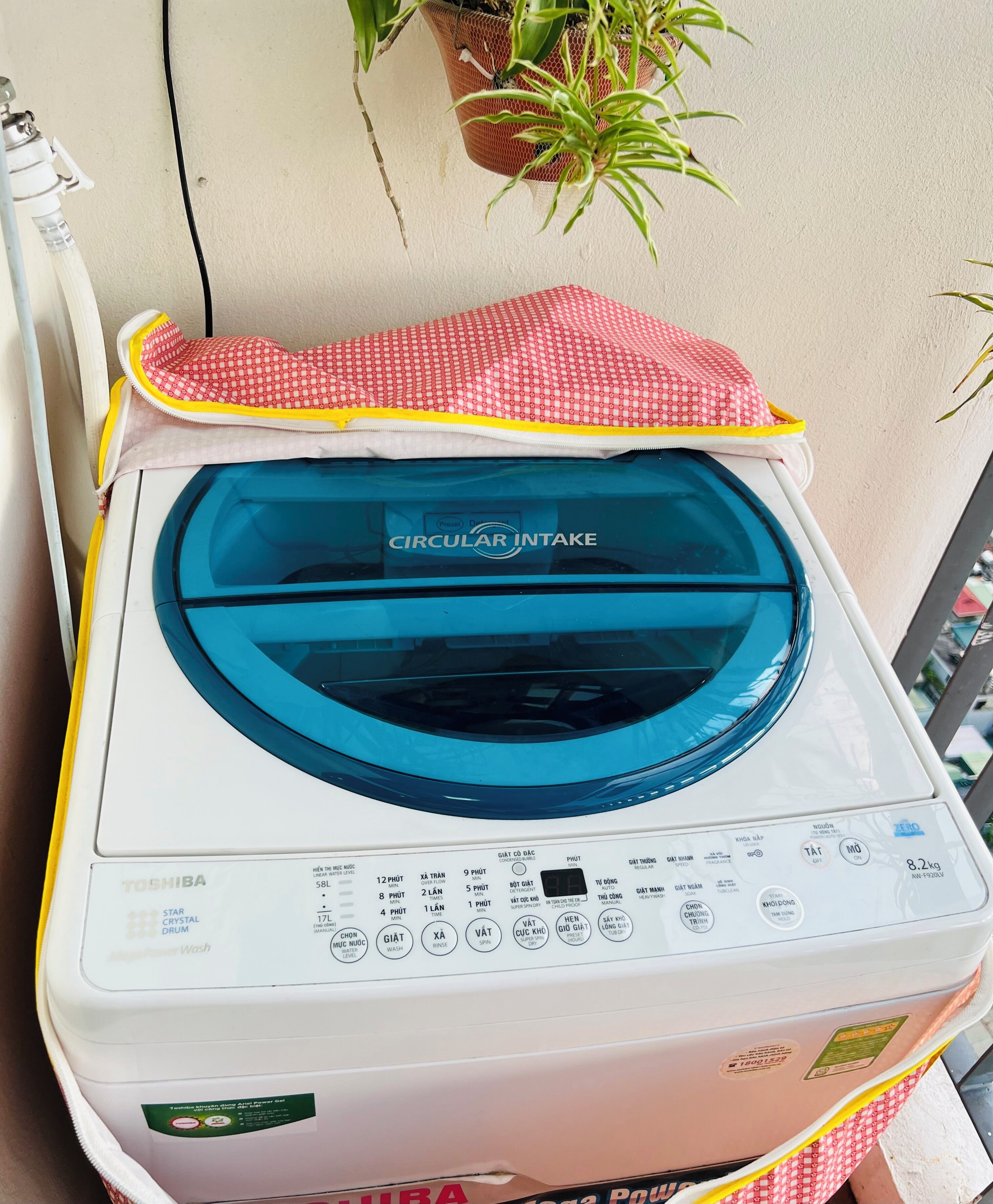 Una lavadora de carga frontal con pantalla digital y varios botones de control, ubicada en un balcón con una planta en maceta y un mantel a cuadros rojo y blanco.