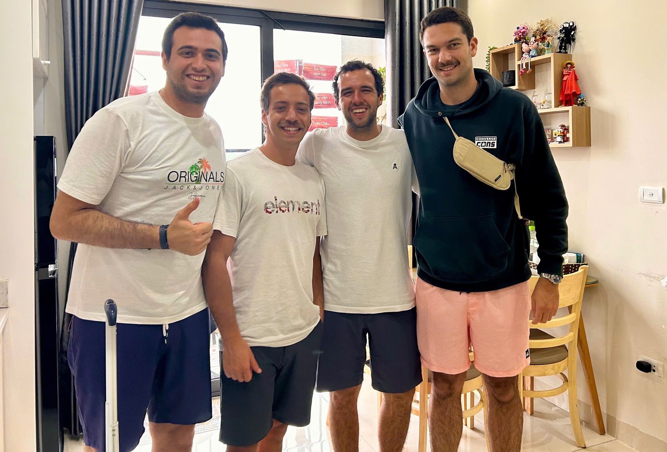 Four men standing in a room with a window, a wooden chair, and a shelf with various items.