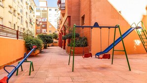 A playground with swings and a slide, surrounded by buildings and a tiled walkway.
