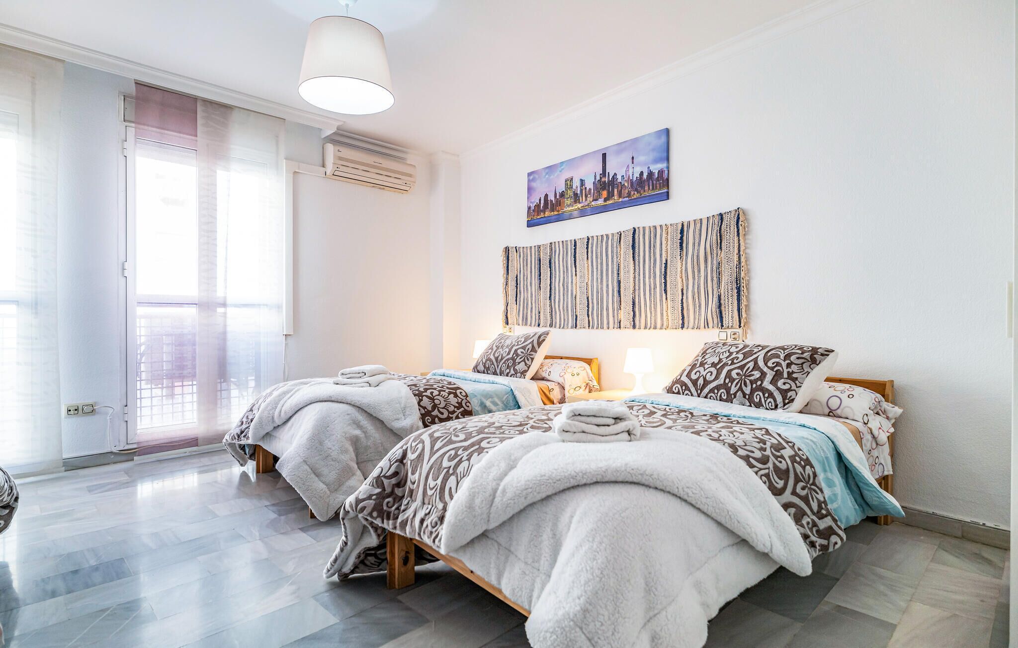 A hotel room with two beds, a cityscape painting, a textured wall hanging, and a large window with a view.