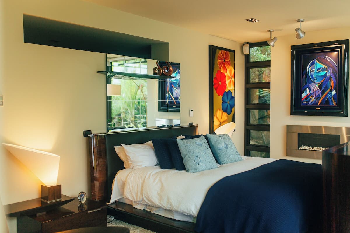 Une chambre à coucher moderne avec un lit, des tables de chevet, un miroir, des œuvres d’art et une cheminée.