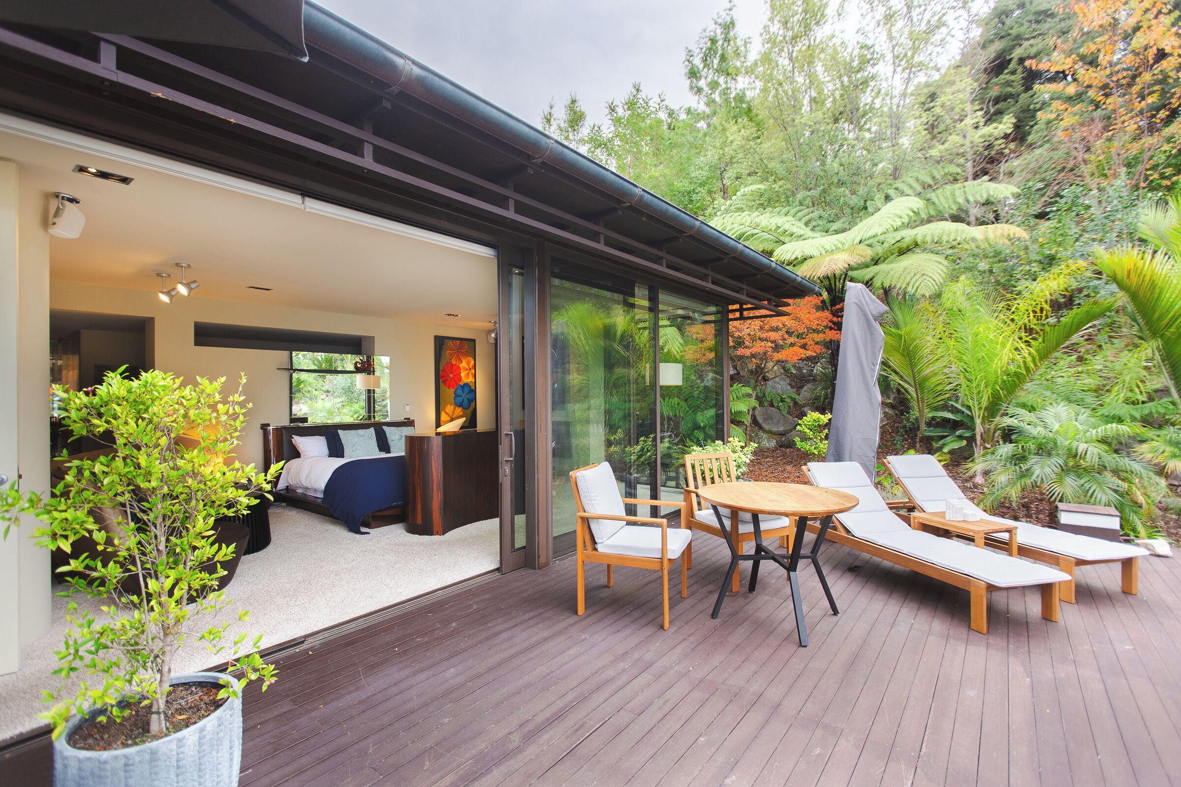 A modern outdoor deck with a wooden table and lounge chairs, surrounded by lush greenery and a glass door leading to a bedroom with a bed and a painting.