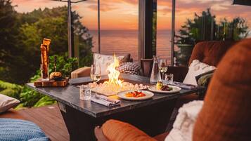 A dining table with a fire, plates of food, and glasses of wine, set against a scenic view.