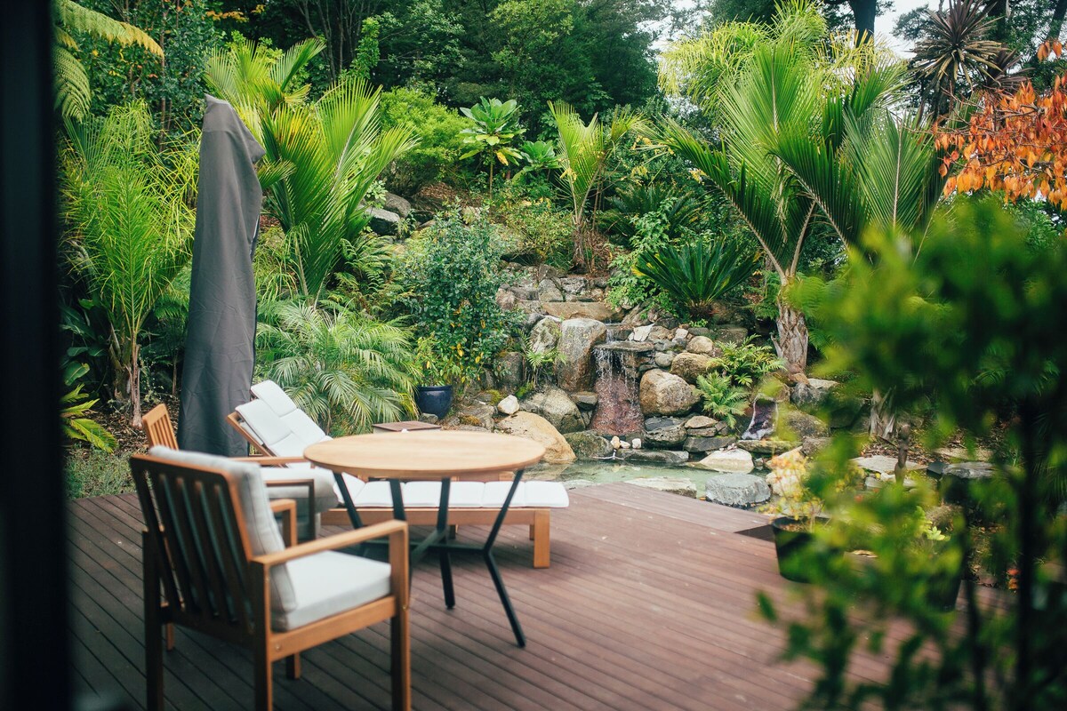 Een houten terras met een tafel en stoelen, omringd door weelderige begroeiing en een kleine waterval.