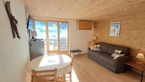 Et hyggeligt rum med træloft, et spisebord med stole, en grå sofa med puder og et vindue med udsigt over det fri.