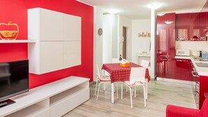 Un salon moderne avec un mur d’accent rouge, un téléviseur à écran plat, une table à manger recouverte d’une nappe à carreaux rouges et noirs, et des chaises blanches.