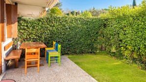 Une terrasse avec une table et des chaises en bois, un chemin de gravier et une haie bien entretenue.