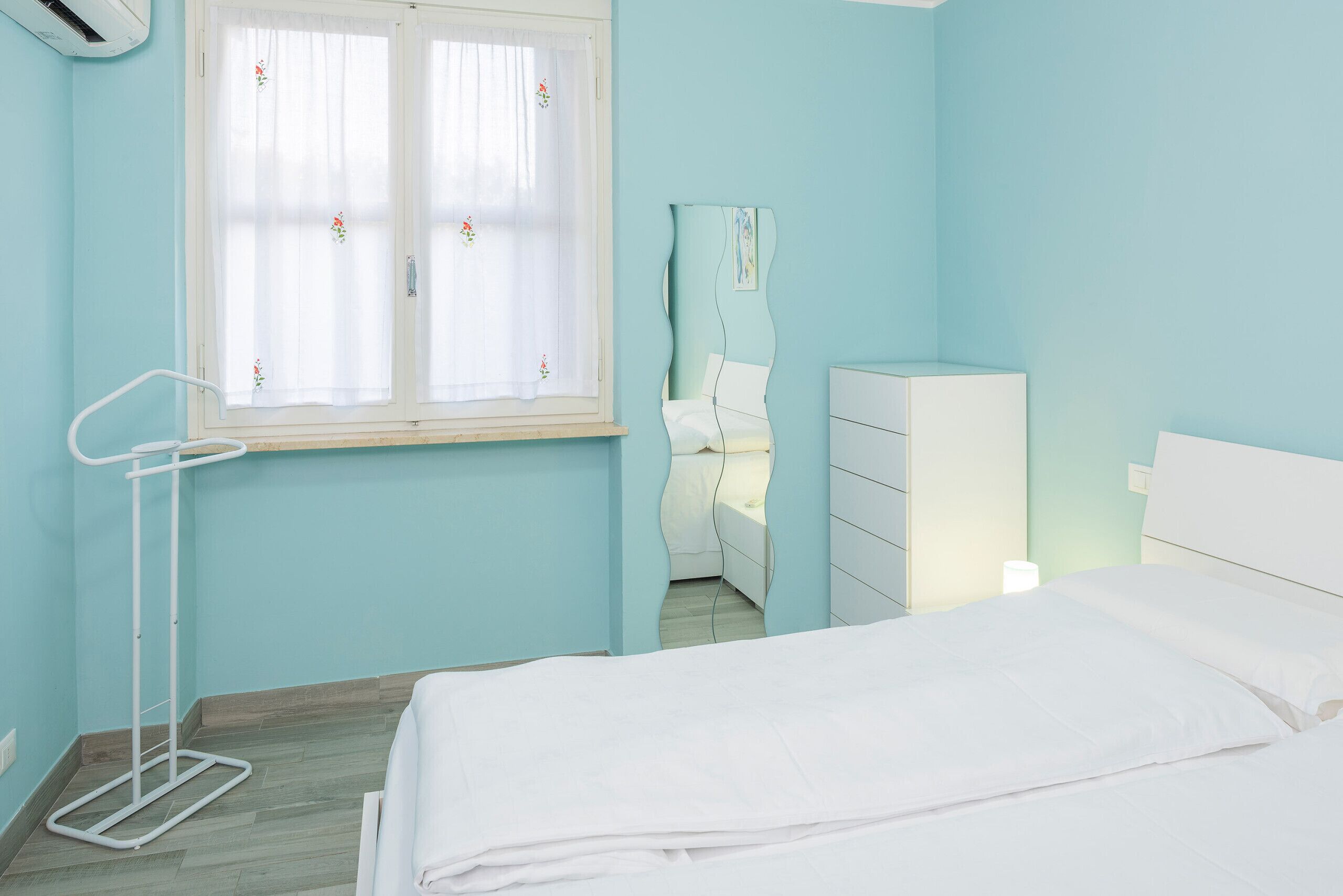 A bedroom with a bed, a dresser, a nightstand, a window with curtains, and a wall-mounted air conditioning unit.