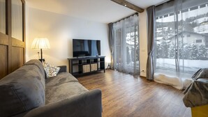 A living room with a sofa, a TV on a stand, a lamp, and a large window with curtains. - 1002A Apartments No.7 Sella (Ortisei)