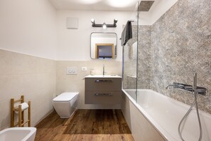 A modern bathroom with a wooden floor, a white toilet, a sink with a mirror above, and a bathtub with a showerhead.