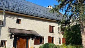 Ein zweistöckiges Gebäude mit einem geneigten Metalldach, Holzläden und einer kleinen Veranda mit zwei Holzstühlen und einer Bank.